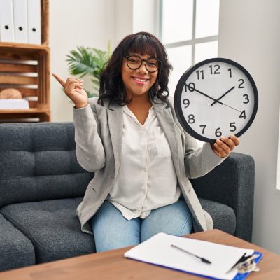 Hispanic woman working at therapy office holding clock smiling happy pointing with hand and finger to the side